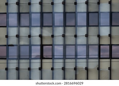 Storm Clouds Reflect in Repeating Office Windows as a Modern Glass Facade Turns the Evening Sky into Urban Geometry - A close-up view of a contemporary building facade reveals a precise grid of windows and translucent glass panels reflecting a dramatic evening sky. Repeating vertical and horizontal structural elements create a strong geometric rhythm across the composition, while subtle reflections of clouds add depth and atmosphere to the otherwise orderly architectural pattern. The combination of muted tones, reflective surfaces, and strict symmetry transforms an ordinary office exterior into a visually striking study of modern design. The photograph explores themes of urban architecture, repetition, structure, and the interaction between the built environment and changing weather conditions.