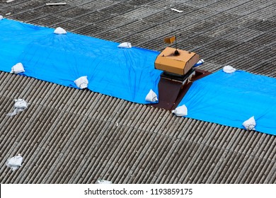 Storm Damaged Roof On House With A Protective Blue Plastic Tarp Spread Over Hole In The Shingles And Rooftop.