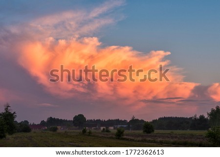 Stormcloud with sunset light on the twilight sky. Huge fluffy cloud in orange and pink colour. Big cumulus clouds over the rural landscape