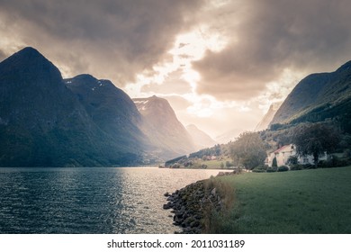 Stormclouds with sun shining through them above dramatic mountain range of Norwegian fjord. Viewed from the shore with white Scandinavian house nearby