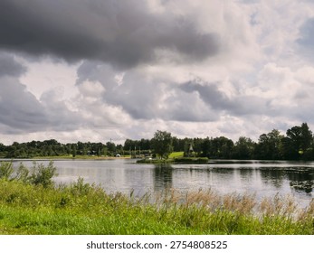 Stormy clouds hover over calm lake waters. Green grasses sway gently in the breeze. Trees line the distant shore under a brooding sky. Water reflects the sky's heavy, textured clouds