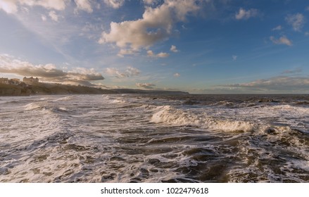 A Stormy Sea With Waves Rushing To The Shore. A Blue, Cloudy Sky Is Overhead.