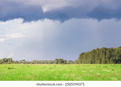 A stormy sky with a large cloud in the distance. The sky is mostly blue with a few white clouds. The grass is green and the trees are tall - Powered by Shutterstock