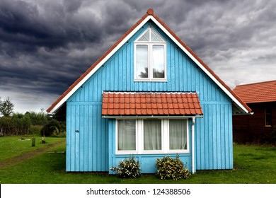 A Stormy Sky Surrounds A Small And Peaceful Blue Wooden House