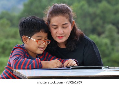 The Story Time Of Aunt And Grandchild. The Asian Woman And Cute Boy Are Reading The Book Together At Table With Blurred Background Of Green Tree