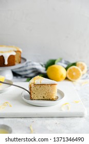 straight ahead shot of a single slice of a lemon sponge cake with cake slice underneath it on a small white plate and some lemons in the background