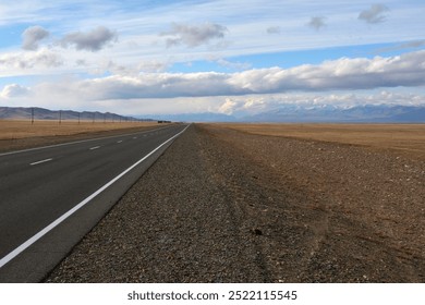 A straight asphalt road goes through the autumn steppe, surrounded by high snow-capped mountains along power line poles. Chuisky tract, Altai, Siberia, Russia. - Powered by Shutterstock