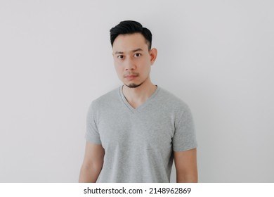 Straight Face Portrait Of Serious Asian Man In Grey T-shirt On White Wall.