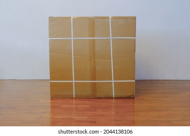 Straight Front View Of Large Brown Blank Cardboard Box On Wooden Floor, Against White Wall. For Advertising Branding Template Mock Up Purpose. Home Delivery Concept.