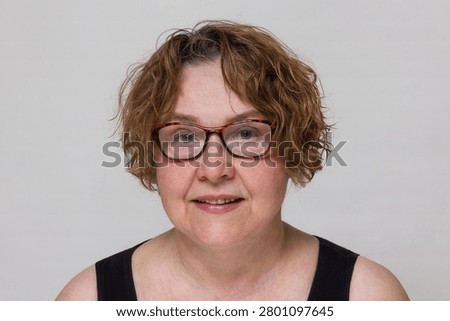 A straight front view studio passport portrait of an authentic mature Caucasian woman with short curly brown hair wearing eyeglasses against a neutral gray background.