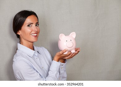 Straight Hair Woman Holding Pink Piggy Bank With Toothy Smile And Formal Clothing While Looking At Camera On Grey Texture Background