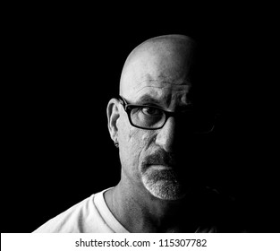 Straight On Black And White Head Shot Of A Middle Aged Man Looking Into Camera With Facial Hair And Glasses With Half His Face Shadowed On A Black Background