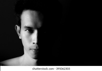 Straight On Black And White Head Shot Of A Man Looking Into Camera With Half His Face Shadowed On A Black Background