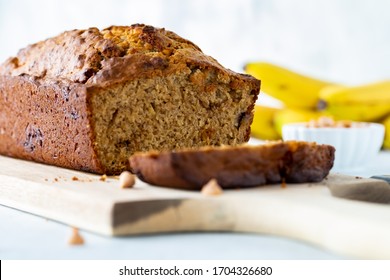 A Straight On Close Up View Of A Banana Bread With One Slice In Front, Resting On A Wooden Board.