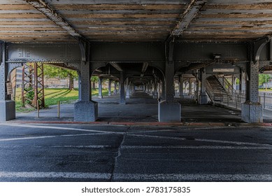 A straight on shot under the elevated Long Island Railroad steel under the Valley Stream, NY train station. In view are empty parking spots during the MTA strike, weekday rush hour. Riveted columns, arched beams, aged patina and dappled natural light. Perfect for editorial, architecture, infrastructure and urban branding.