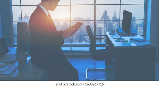 Straight Plane Angle View Of Confident Businessman Working On Digital Tablet While Standing In Front Of The Big Window In Office. The View Outside The Window Is The City. Strategy Business Concept.