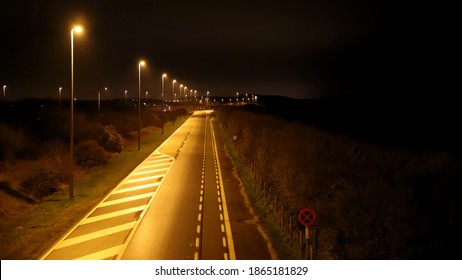 Straight Road In The North Of Belgian Flanders. Large Street Lights Cast A Yellow Light On The Road. Night Photo. An Illuminated City Is Visible In The Background. Way Of Communication. 