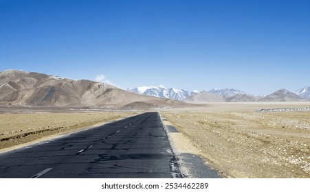 A straight road stretches toward distant snow-capped peaks, surrounded by arid terrain under a bright blue sky. - Powered by Shutterstock