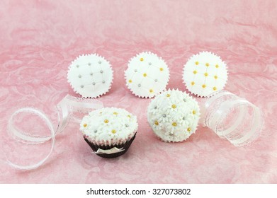 Straight View Small Cupcakes Of Different Designs.  Fondant Topping Decorated With Edible Beads Of Golden And Silver Colors.  Pink Floral Background And Have White Lacy Ribbon As Decoration.