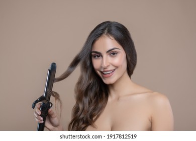 Strand of hair. Beautiful long-haired smiling woman with bare shoulders and expressive look holding lock of hair in curling iron