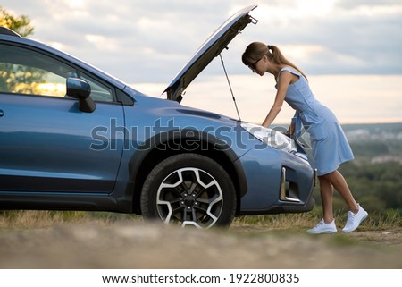 Stranded young woman driver standing near a broken car with popped up bonnet inspecting her vehicle motor.