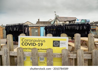 Strandhill Beach, County Sligo, Ireland - 12.07.2021: COVID 19 HSE Poster On A Wooden Fence. Wet Suits Hanging On A Rack In Front Of A Surf School