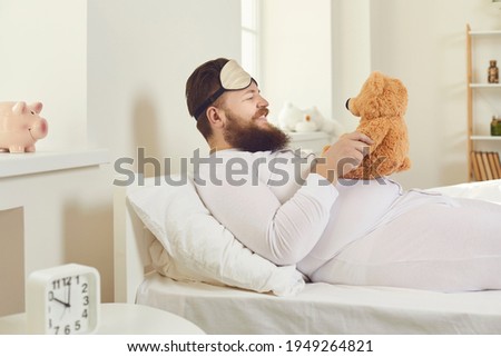 Strange behavior in adults. Side view of a funny bearded overweight man lying in bed with a small teddy bear. Cheerful man in white pajamas and a sleeping mask woke up and behaves like a child.