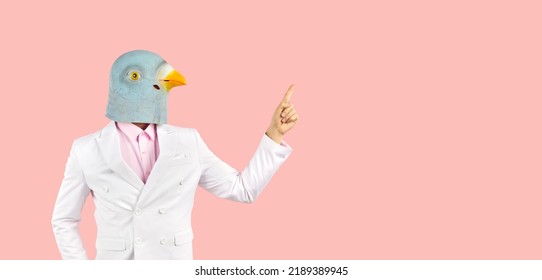 Strange guy with funny bird head points his index finger at blank copy space on right side. Man in suit and wacky pigeon mask showing something on solid pastel pink text copyspace banner background