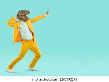 Strange guy in reptile costume dancing and having fun. Crazy young man wearing yellow suit and funny dinosaur mask dancing and moving hands isolated on blank turquoise copy space background