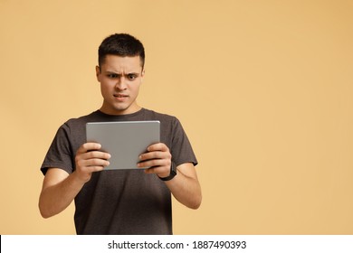 Strange Video, Shocking Information, Weird Message. Surprised And Upset Millennial Man In Brown T-shirt Looks In Digital Tablet And Reads News, Isolated On Beige Background, Studio Shot, Free Space