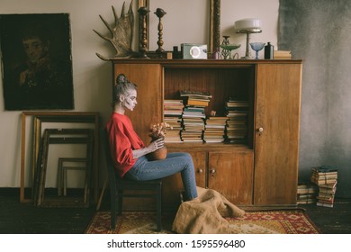 Strange woman wirh conoc stripe body art siiting chair and vase dry flowers in her hands against cupboard and books 