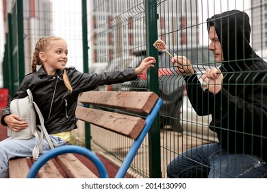 Stranger Offers Candy To Child Sitting On Playground. Kid In Danger. Children Safety Protection Kidnapping Concept. Smiling Happy Caucasian Girl In Leather Jacket Is Affable, Don't Afraid Of Strangers