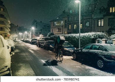 STRASBOURG, FRANCE - DEC 3 2017: Deliveroo Delivery Employee On Bike In French City Cycling Fast For Food Delivery On Time On A Cold Winter Snowy Night In Residential Neighborhood With Cars Parked