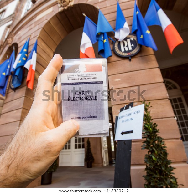 Strasbourg, France - May 26, 2019: Square\
image of man hand holding Voter\'s car French Carte Electorale with\
entrance to Hotel de Ville city hall polling station on the 2019\
European Parliament