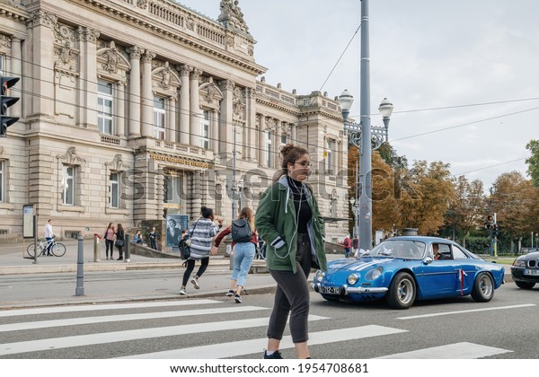 Strasbourg, France- Sep 16, 2017: Woman with\
headphones crossing street in front of luxury vintage sport car\
with The National Theatre of Strasbourg in background on Place de\
la Republique\
Square