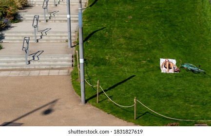 Stratford, London / UK - 13 Sept 2020: Looking Down On An Isolated Girl Sun Bathing In Shorts. She Lies On A White Towel On Grass Beside Wide Empty Steps. Her Shoes Are Off And A Bicycle Nearby.