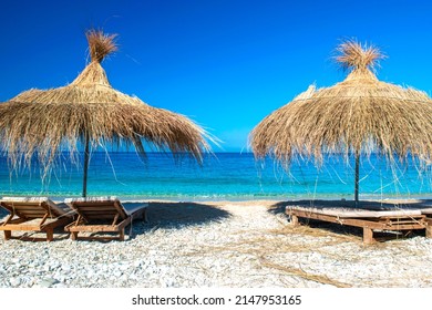 Straw beach umbrellas and comfortable sun loungers on clean sand and pebble beach. Himare. Albania. Ionian Sea. Vacation sea concept. Summer sunny seaside landscape.