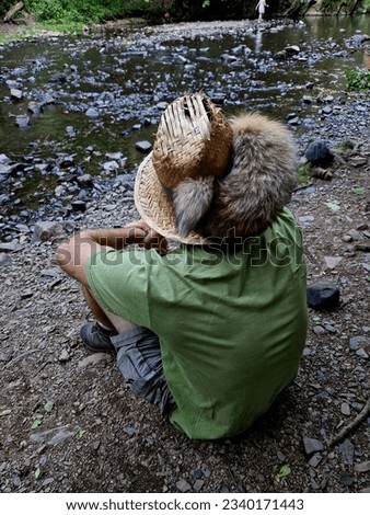 a straw hat with a fox tail is the emblem of hunters and trappers. beer trampers and outdoor devotees take these furs on their hats as decoration. a man sits on the bank of a stream.