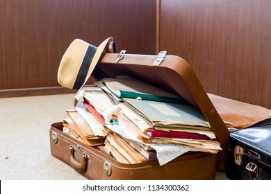 Straw hat is hanging on edge, of old fashioned suitcase made from leather, who is full of folders, papers and documents, overloaded.