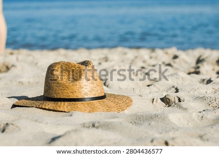 Straw hat placed on sandy beach with clear ocean water in the background and a blurred figure standing nearby under bright sunlight