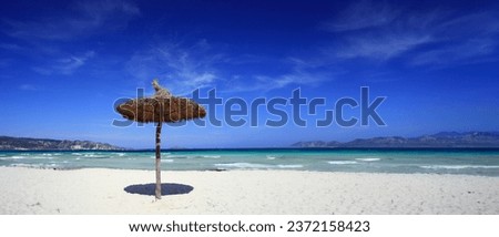 Straw umbrella on Alcudia beach, Palma de Mallorca. Midday sun highlights the clear water, deep blue sky, with distant low mountains on the horizon.

