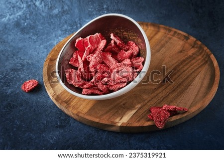 Strawberry chips stacked in ceramic plate on wooden stand on gray-blue concrete background. Hundred-percent natural sublimated strawberries. Healthy vegetable chips