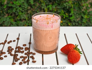 Strawberry latte with fresh strawberry and coffee beans on white wooden table against green leaves with copy space