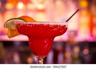 Strawberry Margarita On The Rocks With Salt On Glass Rim And Garnished With Orange, Lemon And Lime Slices Sitting On Bar In Front Of Bar Lights