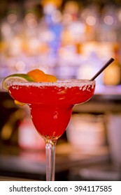 Strawberry Margarita On The Rocks With Salt On Glass Rim And Garnished With Orange, Lemon And Lime Slices Sitting On Bar In Front Of Bar Lights