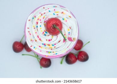 Strawberry Milkshake From Above With Whip Cream, Rainbow Sprinkles, Cherry, On White, With Other Cherries Around The Bottom.