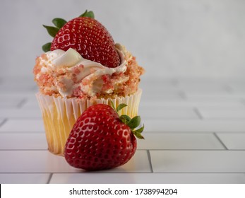 Strawberry Shortcake Cupcake, A Yellow Cupcake Filled With Strawberry Jam, Whipped Cream Swirled High And Strawberry Cake Crumbles Garnished With A Fresh Strawberry On A White Tile Counter.