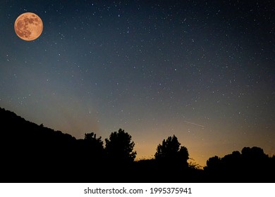 The strawberry supermoon in the dark night in the sky gradient from orange to blue over silhouettes of mountains and trees in June 2020 in Spain.