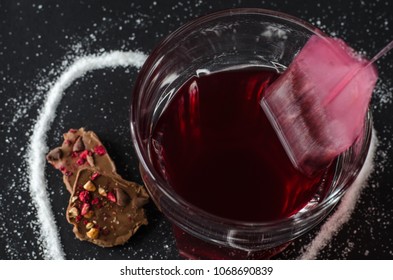 Strawberry Tea And Tea Bag Put Out Of Glass, Spilled Sugar In Shape Of Heart Around, Milk Chocolate, On Dark Background