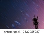 Streaks of vibrant light trails across a twilight sky, with a silhouetted antenna tower on the side.